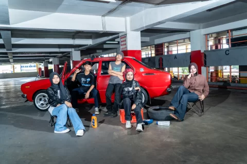 Sekelompok siswa berpose siaga di dekat mobil penjelajah merah di dalam area garasi markas sebelum memulai misi petualangan