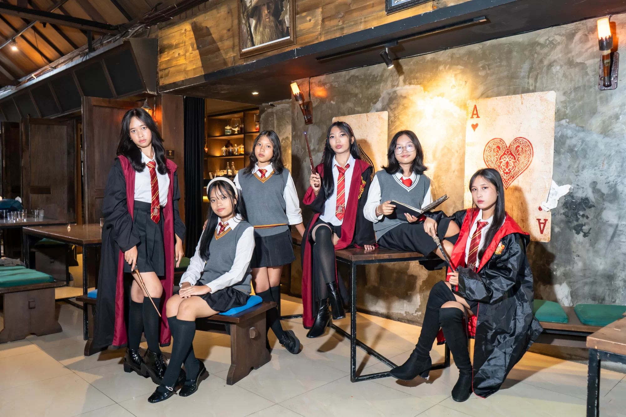 Sekelompok siswa berpose mengenakan jubah asrama Hogwarts dan memegang tongkat sihir di aula besar yang megah
